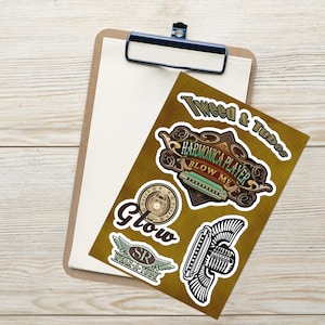 Puede incluir: Un conjunto de pegatinas con diseños de armónica. Las pegatinas incluyen textos como "Tweed & Tubes" y "Harmonica Player Blow Me". La paleta de colores incluye marrón, verde y blanco, con una estética vintage. Las pegatinas están colocadas en una carpeta.