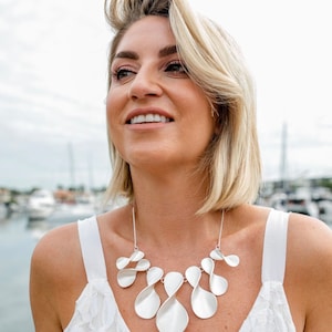 May include: A woman wearing a white lace top with a silver necklace featuring a unique, abstract design. The necklace is made of multiple interconnected, curved, and twisted pieces, resembling petals or leaves.
