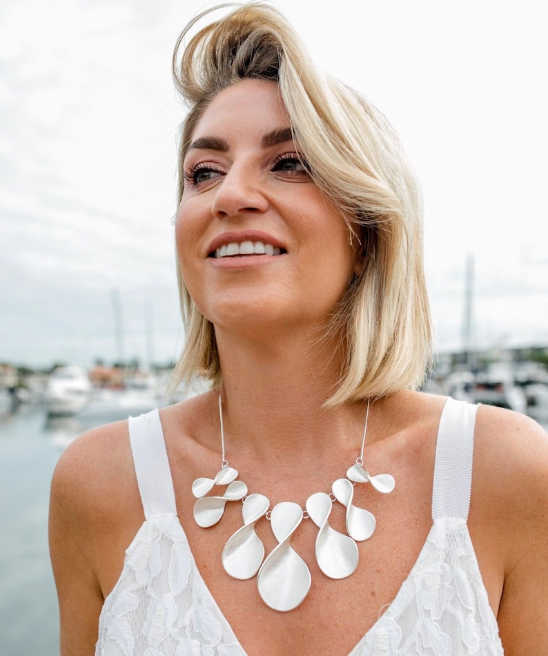 May include: A woman wearing a white lace top with a silver necklace featuring a unique, abstract design. The necklace is made of multiple interconnected, curved, and twisted pieces, resembling petals or leaves.