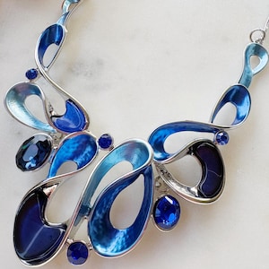 May include: A silver necklace with a fluid design. It features open shapes in various blue shades, from light to navy, and is embellished with small, round, and oval blue gemstones. The necklace is presented on a white surface.