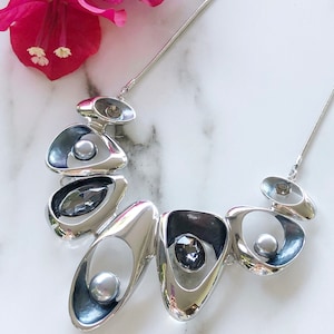 Puede incluir: Un collar de declaración plateado con un diseño geométrico. El collar presenta múltiples formas ovaladas entrelazadas con piedras grises y blancas.
