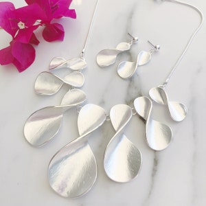 May include: A silver necklace and earrings set featuring a twisted, leaf-shaped design. The necklace has multiple layers of the design, while the earrings have a single layer.