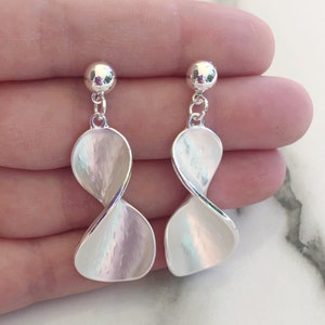 Puede incluir: Un par de pendientes de plata con un diseño retorcido, blanco y nacarado. Los pendientes son de estilo colgante y tienen una pequeña bola de plata en la parte superior.