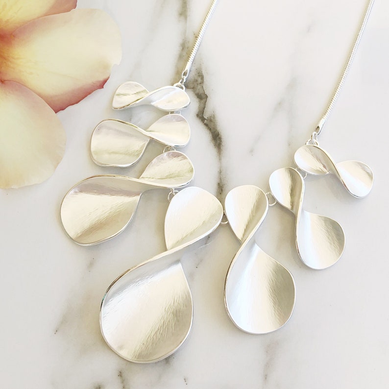 May include: A silver necklace with a layered design featuring multiple twisted, teardrop-shaped pendants. The necklace is on a white marble surface.