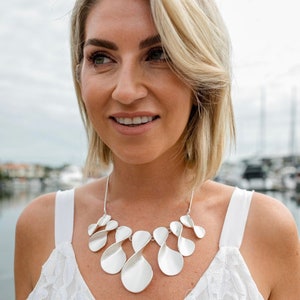 May include: A woman wearing a white lace top with a silver statement necklace featuring a series of interconnected, twisted, and elongated ovals.