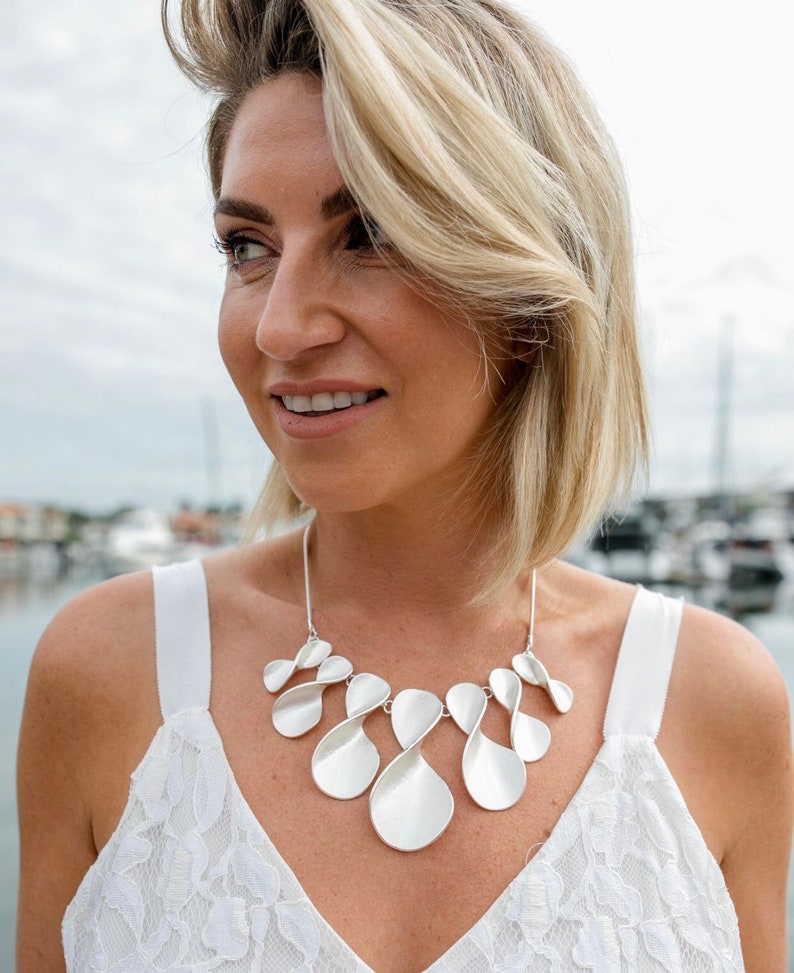 May include: A silver statement necklace with a unique, organic design. The necklace features multiple interconnected, teardrop-shaped pieces that create a flowing, asymmetrical look.