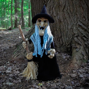 Op de afbeelding: Gehaakte heksenpop met een zwarte hoed, zwarte jurk en blauwe sjaal, die een bezem vasthoudt. De pop heeft een lachend gezicht en staat tegen een bosachtergrond met een grote boom. Halloween decoratie.