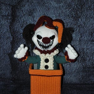 May include: Crocheted jack-in-the-box toy featuring a scary clown with a white face, black eyes, and a menacing grin. The clown wears a green and red outfit with a ruffled collar and a jester hat. The box is orange.