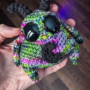 May include: A crocheted stuffed toy with large black eyes, black yarn horns, and a black yarn nose. The toy is made with a variety of colorful yarns, including green, pink, and purple.