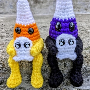 May include: Two crocheted candy corn characters with white ghost figures in their arms. The candy corn character on the left is yellow, orange, and white. The candy corn character on the right is purple, white, and black.