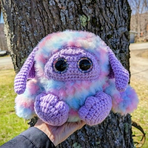 Puede incluir: Un juguete de peluche tejido a crochet con un cuerpo morado, pelaje rosa, azul y blanco, y grandes ojos negros. El juguete tiene una textura esponjosa y se sostiene en una mano.