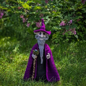 Può includere: Una bambola da mago all'uncinetto che indossa un cappello a punta viola e un mantello viola abbinato sopra una veste nera. La figura ha una lunga barba grigia, un viso bianco e tiene un bastone di legno marrone con una sfera verde scuro.
