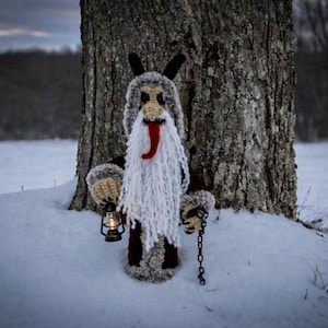 May include: A handmade Krampus doll, crafted with crochet, stands in the snow next to a tree. The doll features a long white beard, red tongue, and black horns. It holds a lantern and a chain.