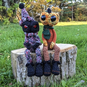 May include: Two handmade crochet dolls sit on a tree stump. One is black with a patterned hat and scarf, red eyes, and black boots. The other is orange with a pumpkin head, large black eyes, a green scarf, and brown legs.