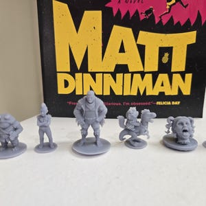 Puede incluir: Una colección de figuras en miniatura grises, cada una con diseños únicos, se organiza frente a un libro titulado "Matt Dinniman". Las figuras incluyen una criatura parecida a una rana, una persona, una figura musculosa, una criatura montada en un dinosaurio, una cabeza gritando y una figura de cabra.