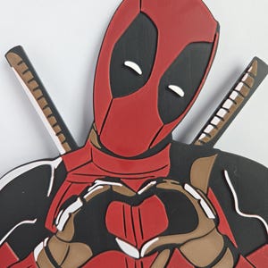 May include: A red, black, and brown Deadpool figure with a heart-shaped hand gesture. The figure has two swords behind its back. The figure is wearing a red suit with black accents and a mask with white eyes.