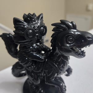 May include: A glossy black figurine depicting a creature riding a dinosaur-like beast. The rider has large goggles and a crown. The dinosaur has detailed scales and a fierce expression. The figurine is mounted on a circular base.