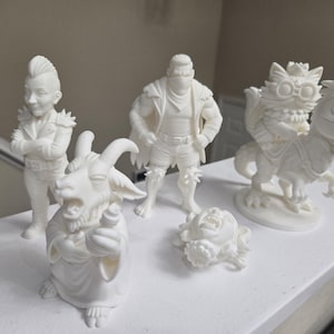May include: A collection of white figurines, including a goat-like creature, a figure with a mohawk, a masked figure, a cat riding a dinosaur, and a small head. The figurines are displayed on a white surface.