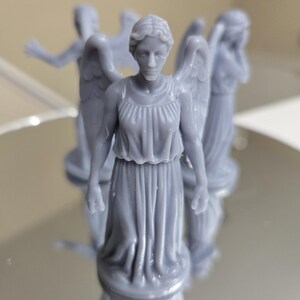 May include: Several grey miniature statues of winged figures, each wearing a flowing gown. The statues are on a reflective surface, with some in focus and others blurred. The figures have detailed facial features and wings.