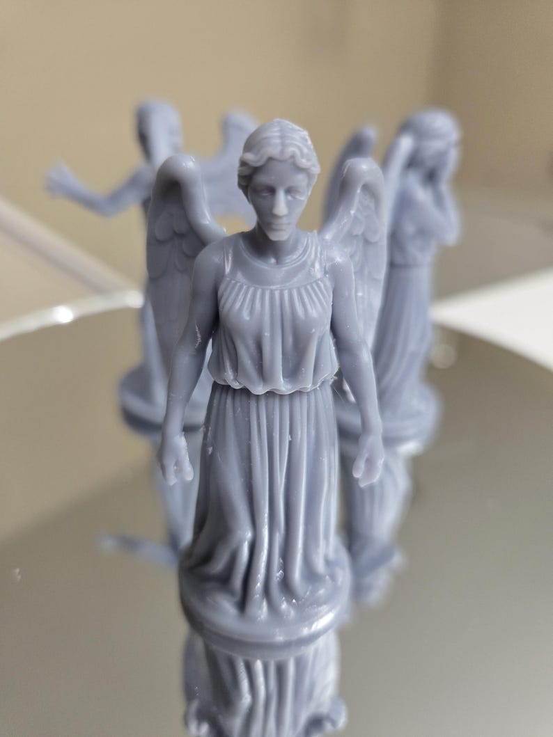 May include: Several grey miniature statues of winged figures, each wearing a flowing gown. The statues are on a reflective surface, with some in focus and others blurred. The figures have detailed facial features and wings.