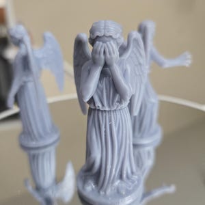 May include: Three pale blue statues of weeping angels, with detailed wings and draped gowns. One angel covers its eyes with its hands. The statues are on a reflective surface, with their reflections visible.