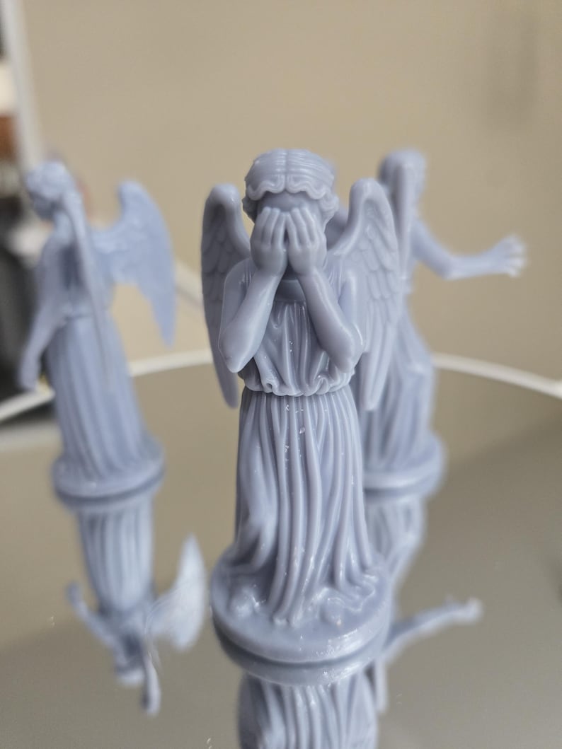 May include: Three pale blue statues of weeping angels, with detailed wings and draped gowns. One angel covers its eyes with its hands. The statues are on a reflective surface, with their reflections visible.