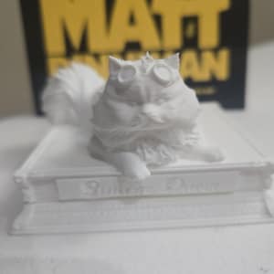 May include: A white 3D-printed cat figurine wearing goggles, resting on a book. The book has the text "Butter's Doom" on the spine. The background features the text "MATT" in large yellow letters.