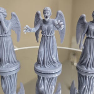 May include: Three grey statues of winged figures in flowing robes. One figure covers its eyes, another has an open mouth and outstretched arms, and the third stands with arms at its sides. The statues are reflected on a shiny surface.
