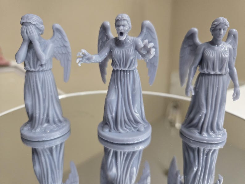 May include: Three grey statues of winged figures in flowing robes. One figure covers its eyes, another has an open mouth and outstretched arms, and the third stands with arms at its sides. The statues are reflected on a shiny surface.