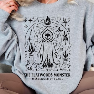 May include: A light gray sweatshirt featuring a black graphic of the Flatwoods Monster, surrounded by flames, candles, and symbols. The text "THE FLATWOODS MONSTER - MESSENGER OF FLAME" is printed below the graphic.