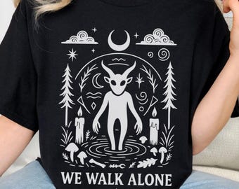 May include: Black t-shirt with a white graphic of a horned figure in a forest scene, surrounded by candles, mushrooms, and a crescent moon. The text "WE WALK ALONE" is printed below the graphic.
