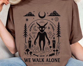 May include: Brown t-shirt with a black graphic design featuring a horned figure in a forest scene, surrounded by candles, mushrooms, and the text "WE WALK ALONE". The design includes a crescent moon and stars.