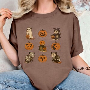 K&ouml;nnte beinhalten: Espressofarbenes T-Shirt mit Halloween-Illustrationen. Das Design zeigt einen Geist, K&uuml;rbisse, ein Hexenopossum und andere gruselige Kreaturen. Das Shirt hat eine bequeme Passform und ist perfekt f&uuml;r die Herbstsaison.