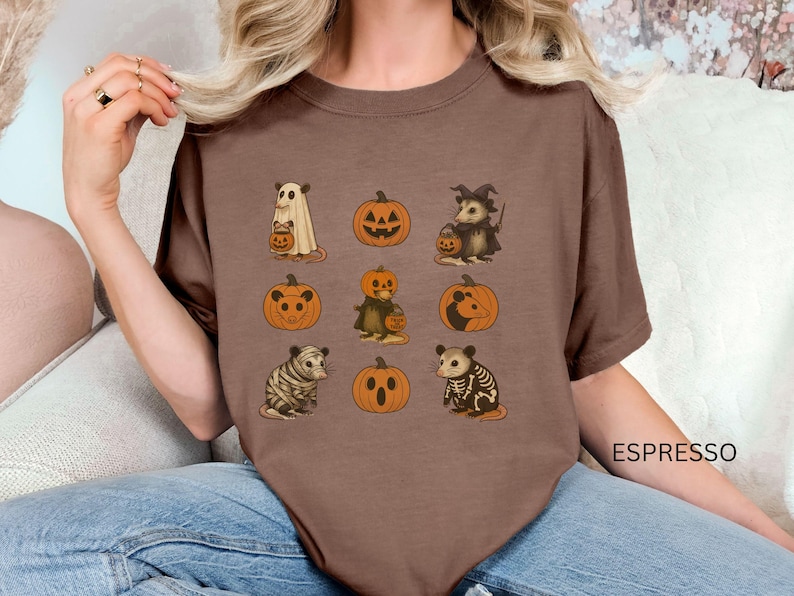 K&ouml;nnte beinhalten: Espressofarbenes T-Shirt mit Halloween-Illustrationen. Das Design zeigt einen Geist, K&uuml;rbisse, ein Hexenopossum und andere gruselige Kreaturen. Das Shirt hat eine bequeme Passform und ist perfekt f&uuml;r die Herbstsaison.