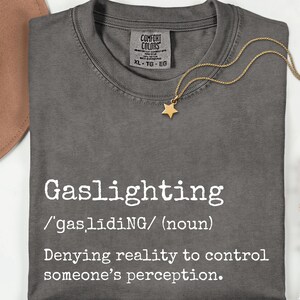 May include: A dark gray t-shirt with the word "Gaslighting" printed in white, along with its definition. A gold star pendant necklace and a brown hat are also visible. The shirt is XL size.