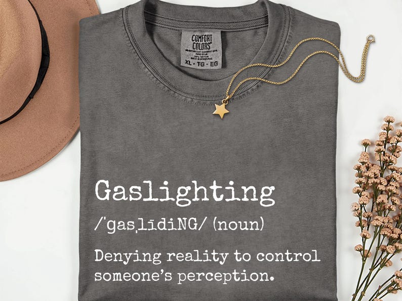 May include: A dark gray t-shirt with the word "Gaslighting" printed in white, along with its definition. A gold star pendant necklace and a brown hat are also visible. The shirt is XL size.