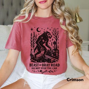 Op de afbeelding: Karmizijnrood T-shirt met een zwarte afbeelding van een weerwolf in een bos, een maan en sterren. De tekst "BEAST OF BRAY ROAD DO NOT STOP THE CAR" staat eronder. Het shirt heeft korte mouwen en een ronde hals.