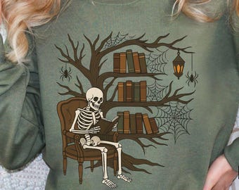 Pode incluir: Camisola verde-oliva com um esqueleto lendo um livro. O design inclui uma árvore com estantes, livros, teias de aranha, uma lanterna e aranhas. Um design único para os amantes de livros.
