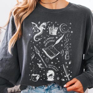 Op de afbeelding: Donkergrijze sweatshirt met een wit grafisch ontwerp met een draak, een kroon, een halve maan, een open boek, een pen en andere mystieke elementen. De tekst "INK IS ANOTHER KIND OF MAGIC" en "MAGIC LIVES IN THE QUIET MOMENTS" zijn ook opgenomen.