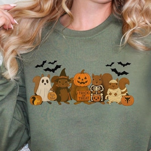 May include: Olive green sweatshirt featuring a Halloween-themed graphic. The design includes cartoon squirrels dressed as a ghost, witch, mummy, and skeleton, along with pumpkins and bats. The words "Trick or Treat" are also present.