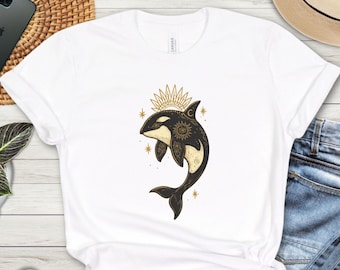May include: White t-shirt with a black and cream orca design, adorned with a sun and moon crown. The orca has detailed patterns and gold stars. The shirt is made of a soft material.