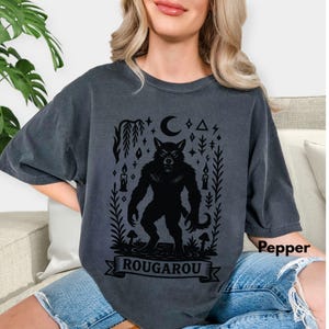 May include: Dark gray t-shirt featuring a black Rougarou graphic. The design includes a werewolf, moon, stars, candles, and the word "ROUGAROU" on a banner. The shirt is a comfortable, oversized fit.
