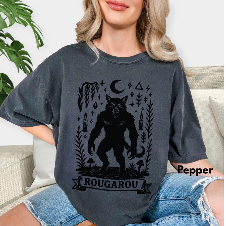 May include: Dark gray t-shirt featuring a black Rougarou graphic. The design includes a werewolf, moon, stars, candles, and the word "ROUGAROU" on a banner. The shirt is a comfortable, oversized fit.