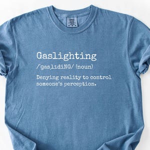 May include: A blue t-shirt with the word "Gaslighting" in white, along with its pronunciation and definition: "Denying reality to control someone's perception."