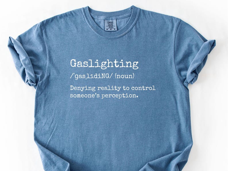 May include: A blue t-shirt with the word "Gaslighting" in white, along with its pronunciation and definition: "Denying reality to control someone's perception."