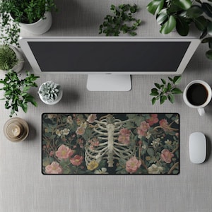 Puede incluir: Alfombrilla de ratón rectangular con una ilustración detallada de una caja torácica humana entrelazada con flores rosas, blancas y amarillas sobre un fondo verde oscuro. La alfombrilla está rodeada por un borde negro.