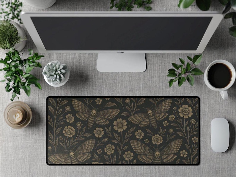 May include: A black desk mat featuring a repeating pattern of moths and floral elements in brown and gold tones. The mat is rectangular and positioned under a computer monitor, with a white mouse and coffee cup. The design has a gothic and nature-inspired aesthetic.