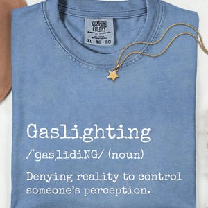 Puede incluir: Una camiseta azul claro con la palabra "Gaslighting" y su definición en blanco. Un collar con un colgante de estrella dorada descansa sobre la camiseta. La camiseta está doblada y se ve parcialmente un sombrero marrón.