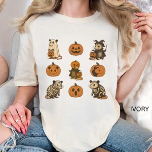 K&ouml;nnte beinhalten: Elfenbeinfarbenes T-Shirt mit Halloween-Illustrationen. Das Design umfasst K&uuml;rbisse und Opossums in Kost&uuml;men, wie ein Geist, eine Hexe und ein Skelett. Das Wort "IVORY" ist auf dem Shirt aufgedruckt.