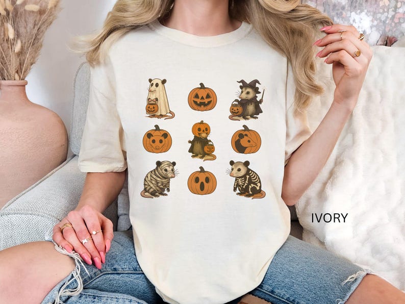 K&ouml;nnte beinhalten: Elfenbeinfarbenes T-Shirt mit Halloween-Illustrationen. Das Design umfasst K&uuml;rbisse und Opossums in Kost&uuml;men, wie ein Geist, eine Hexe und ein Skelett. Das Wort "IVORY" ist auf dem Shirt aufgedruckt.